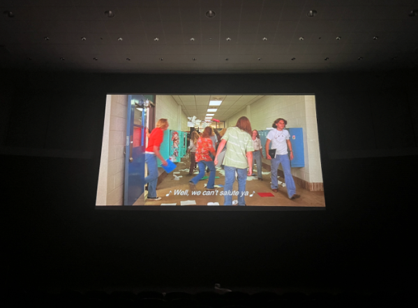 A movie screen in a dark theater depicts teenagers in a high school hallway